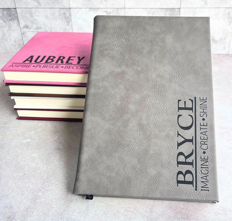 Custom laser-engraved leatherette journals for graduating seniors. Featured in gray and pink with personalized names like 'Bryce' and 'Aubrey' alongside inspirational mottos. A high-quality, permanent graduation keepsake for schools and students.